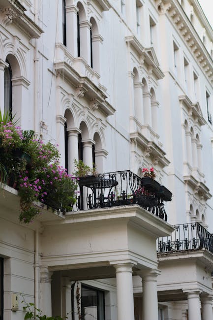 The image depicts the exterior of a white, multi-story building with ornate architectural details, including decorative cornices and window mouldings. Small balconies with black wrought iron railings